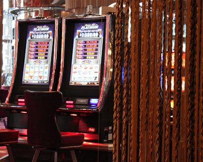 A pair of casino slot machines, each with detailed screens displaying various game outcomes and jackpot amounts. The chairs in front of the slot machines are dark and cushioned for comfort. The background features a hanging curtain made of golden ropes and other casino decorations are visible.