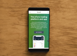 A smartphone displaying an advertisement for an online trading platform called eToro. The screen shows a green background with text promoting the platform as a leading social trading platform. A button labeled 'Get Started' is visible, along with a partial view of a mobile app screen below.