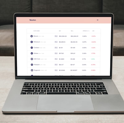 A laptop on a table displaying a cryptocurrency trading platform. The screen lists various cryptocurrencies along with their buy and sell values, spread percentages, and 24-hour change percentages. The keyboard and touchpad of the laptop are visible, set against a neutral background with a sofa in the back.
