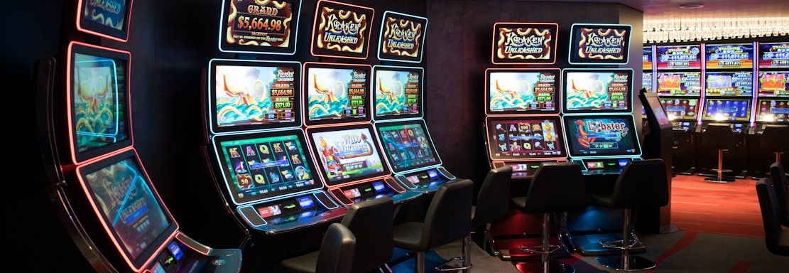 A row of brightly lit slot machines in a casino setting, each with colorful displays and various game themes. The machines are lined up against a dark wall and are unoccupied, with cushioned chairs placed in front of them. The area is dimly lit except for the glow from the screens, creating a vibrant and engaging atmosphere.